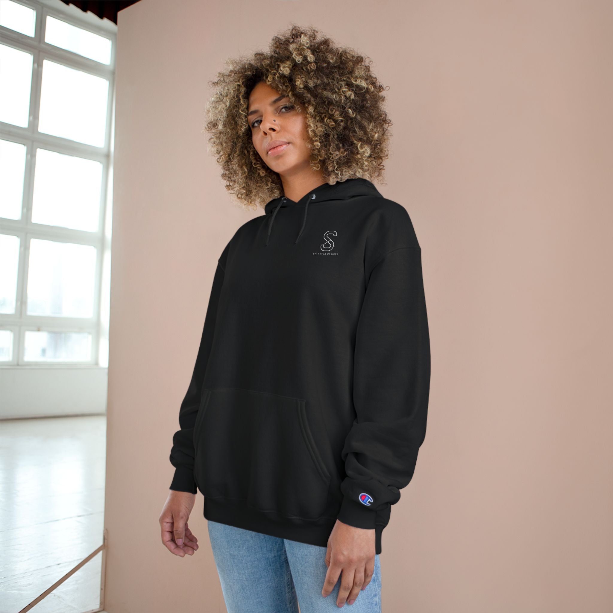 Woman wearing the black SparkyCA Champion hoodie beside a sunlit window