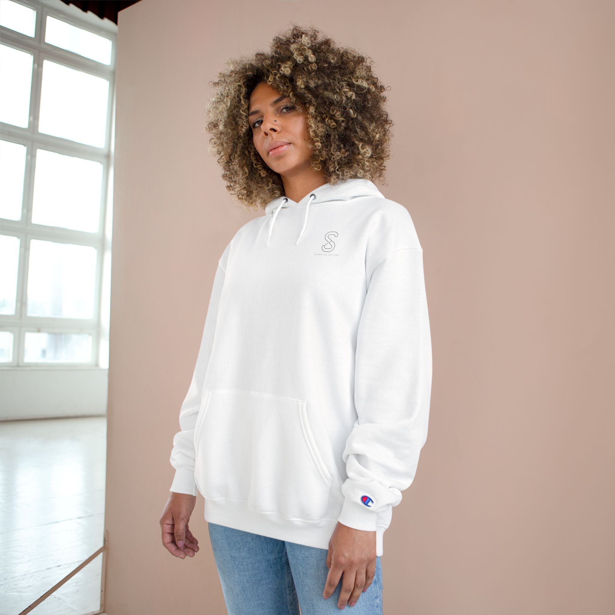 Woman wearing the white SparkyCA Champion hoodie beside a sunlit window