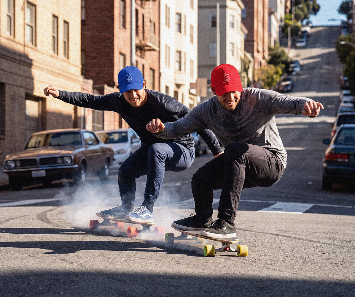 Two skateboarders riding longboards down a steep city street lined with brick buildings. The rider on the left wears a blue cap and dark long-sleeve top, while the rider on the right wears a red cap and gray long-sleeve top.