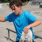Man wearing a blue SparkyCA Designs tee holding a skateboard at a skate park