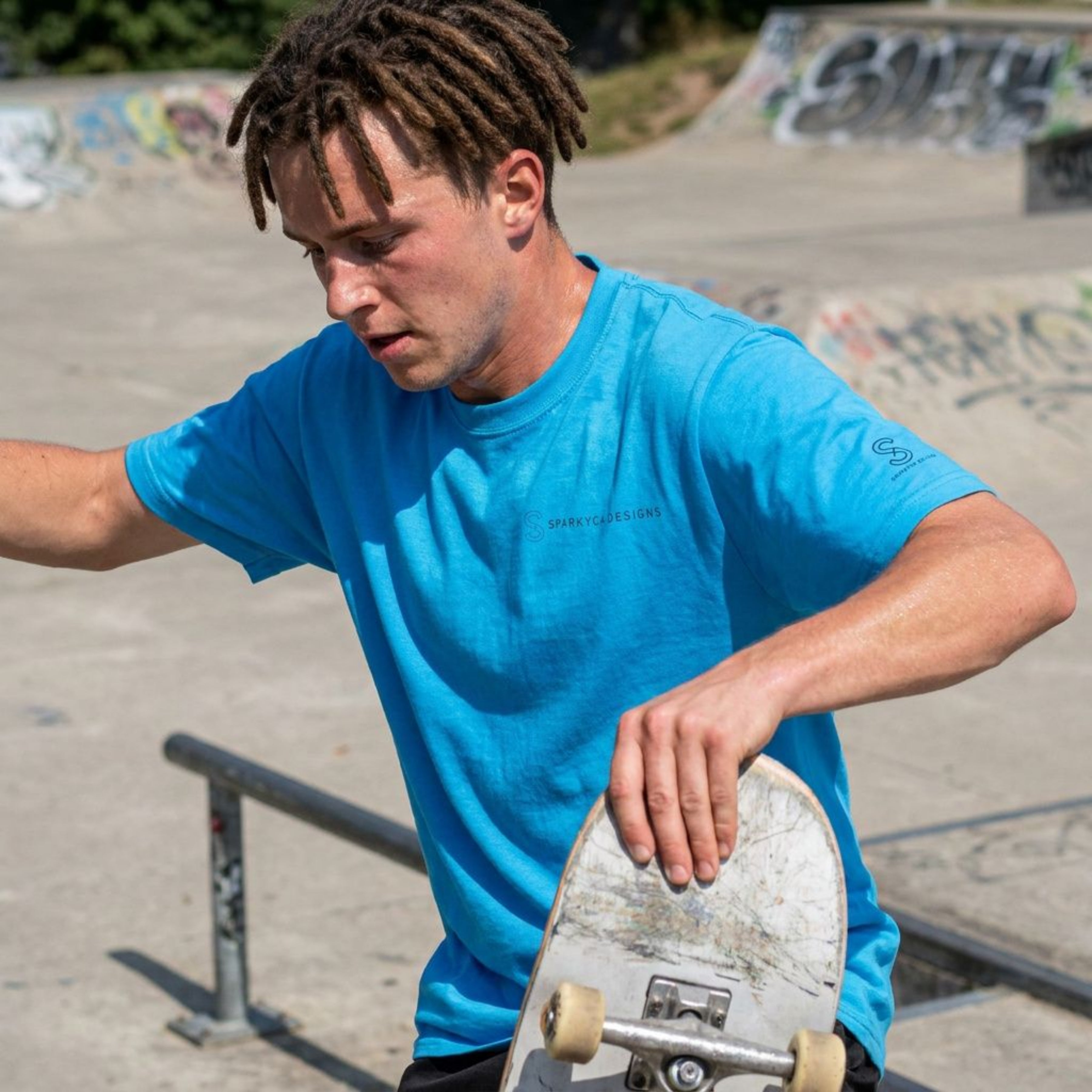Man wearing a blue SparkyCA Designs tee holding a skateboard at a skate park