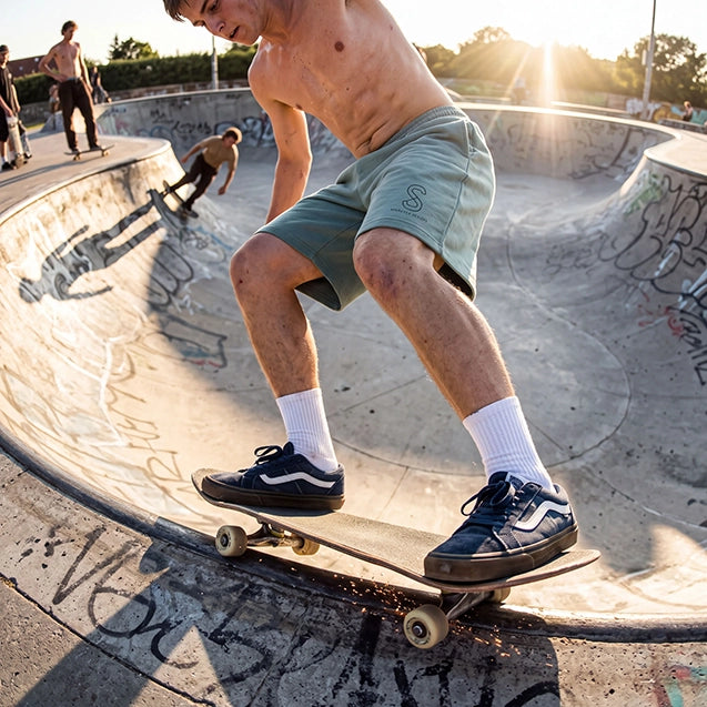 A shirtless skateboarder in sage green shorts, white socks, and dark blue Vans skates along the edge of a concrete bowl at a skatepark during golden hour.